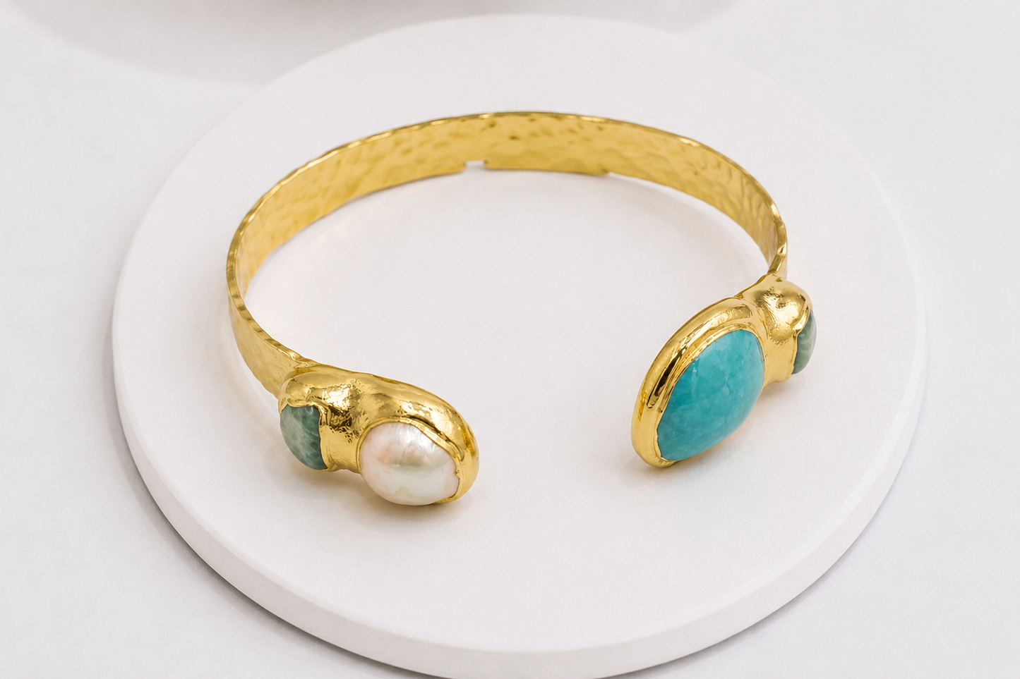 A gold cuff bracelet with a hammered finish, featuring one end with a white pearl and the other with a turquoise stone, displayed on a white circular pedestal.