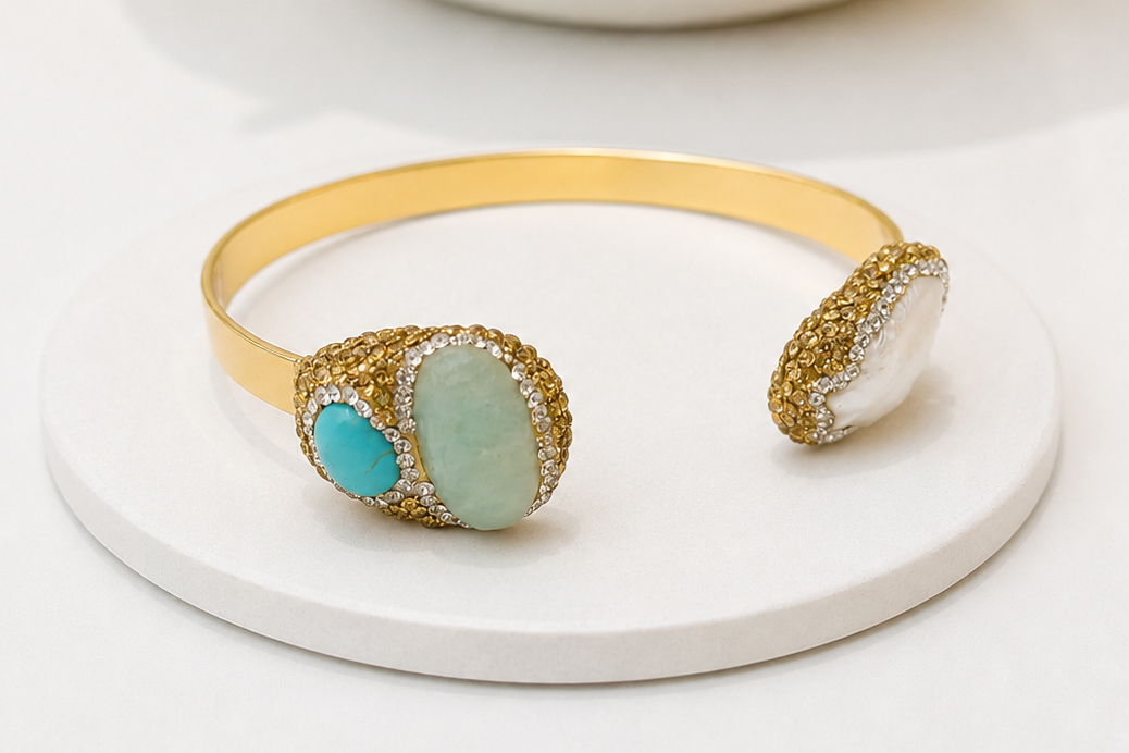 Gold open cuff bangle bracelet with three large oval stone settings, one turquoise and amazonite, on the other side freshwater pearl with sparkling crystal detailing all around on a white background displayed on a round tray. 