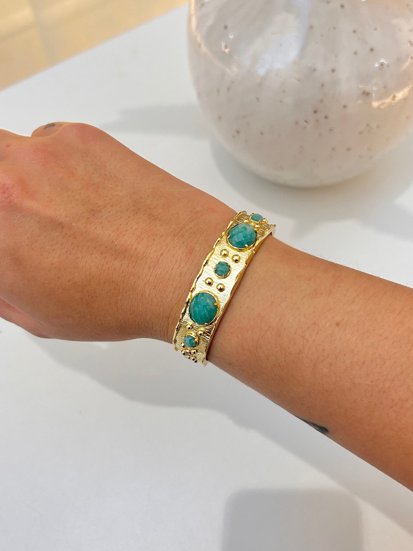 A gold-toned bangle bracelet with round amazonite stones, worn on a person's wrist, with a white table and a speckled ceramic vase in the background.
