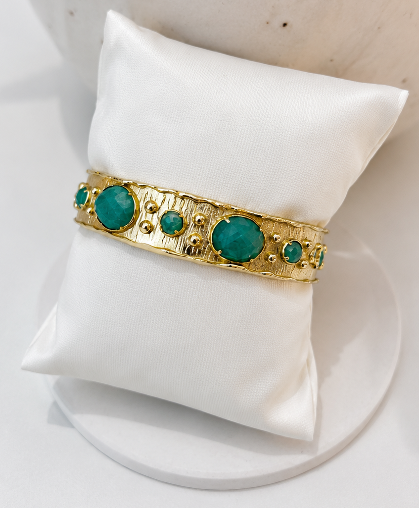 
                  
                    A gold bangle bracelet with large and small round amazonite stones, displayed on a white satin jewelry pillow.
                  
                