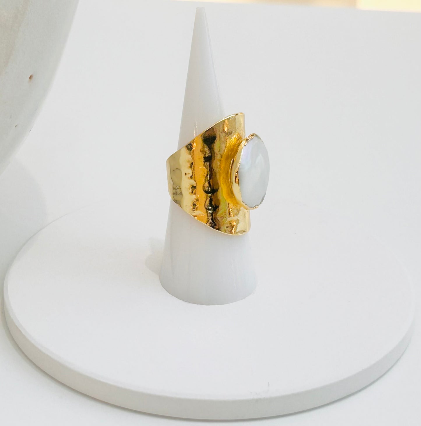 
                  
                    Aureline Freshwater Pearl Statement Ring displayed on a white jewellery stand, featuring a natural pearl set in a wide hammered gold-plated band.
                  
                