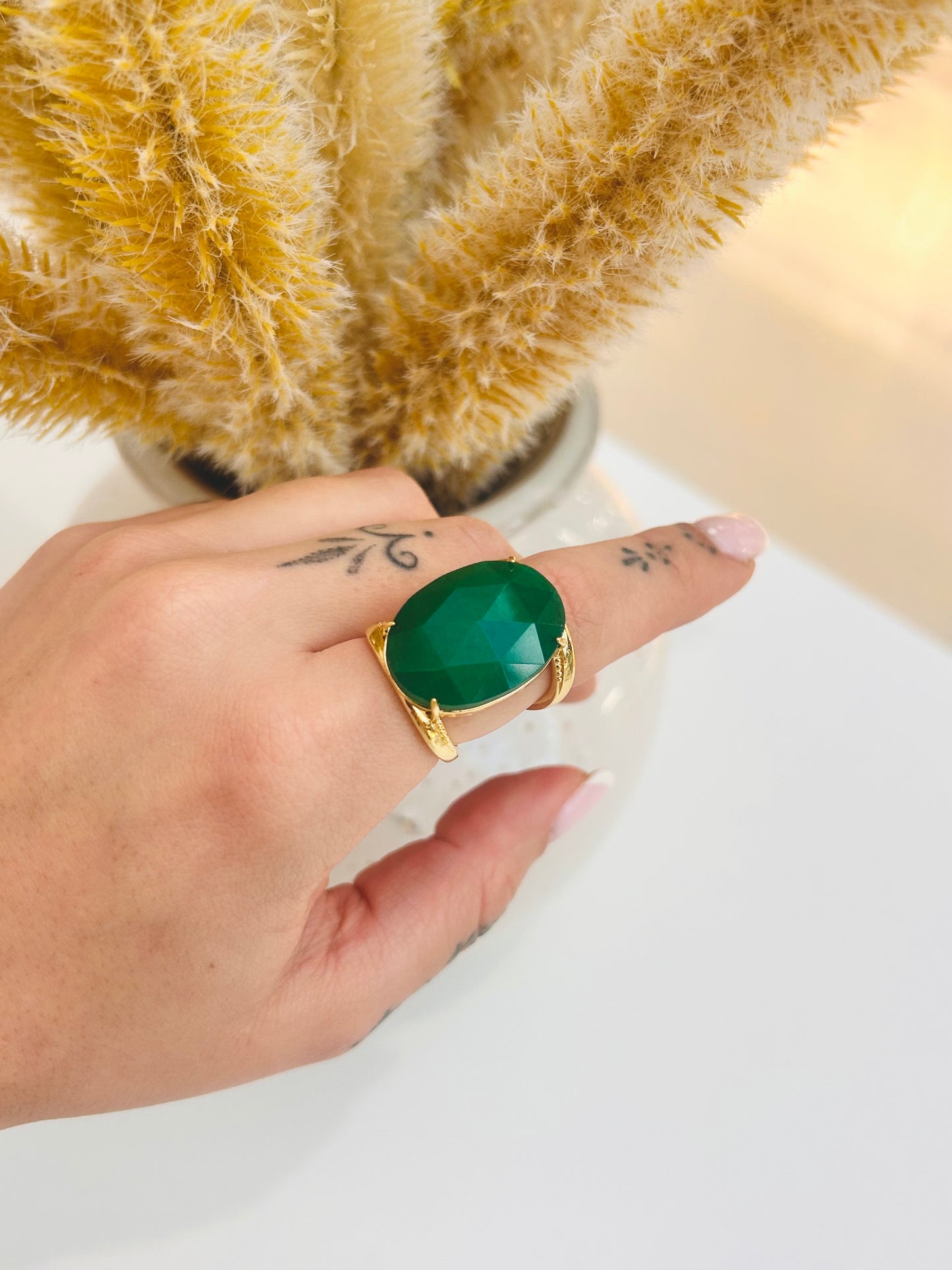 A gold ring with a large oval green jade stone worn on a person's finger, shown against a light background with dried grass in a vase.