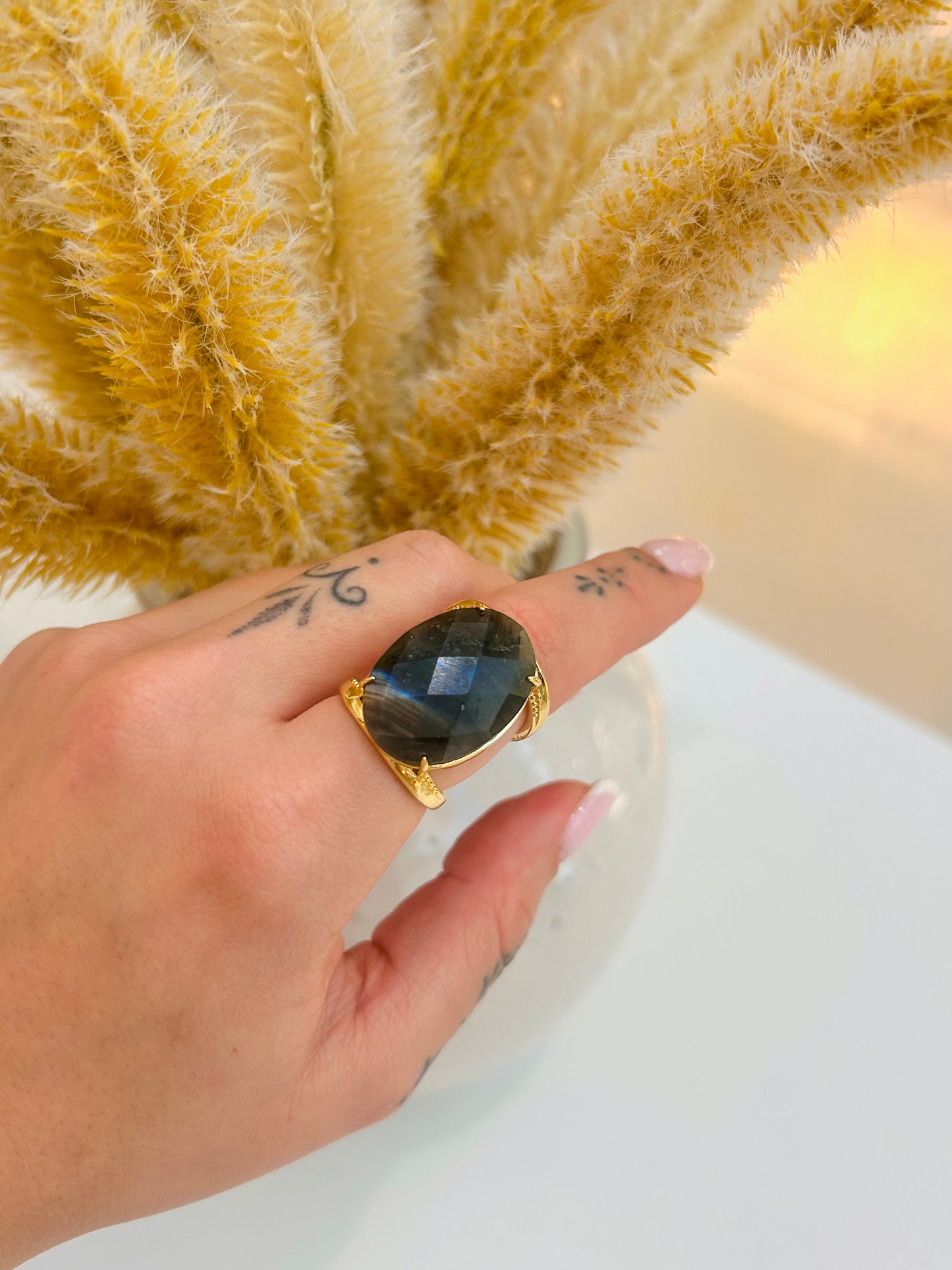A gold ring with a large oval faceted dark labradorite gemstone worn on a person's hand, with dried pampas grass in the background.