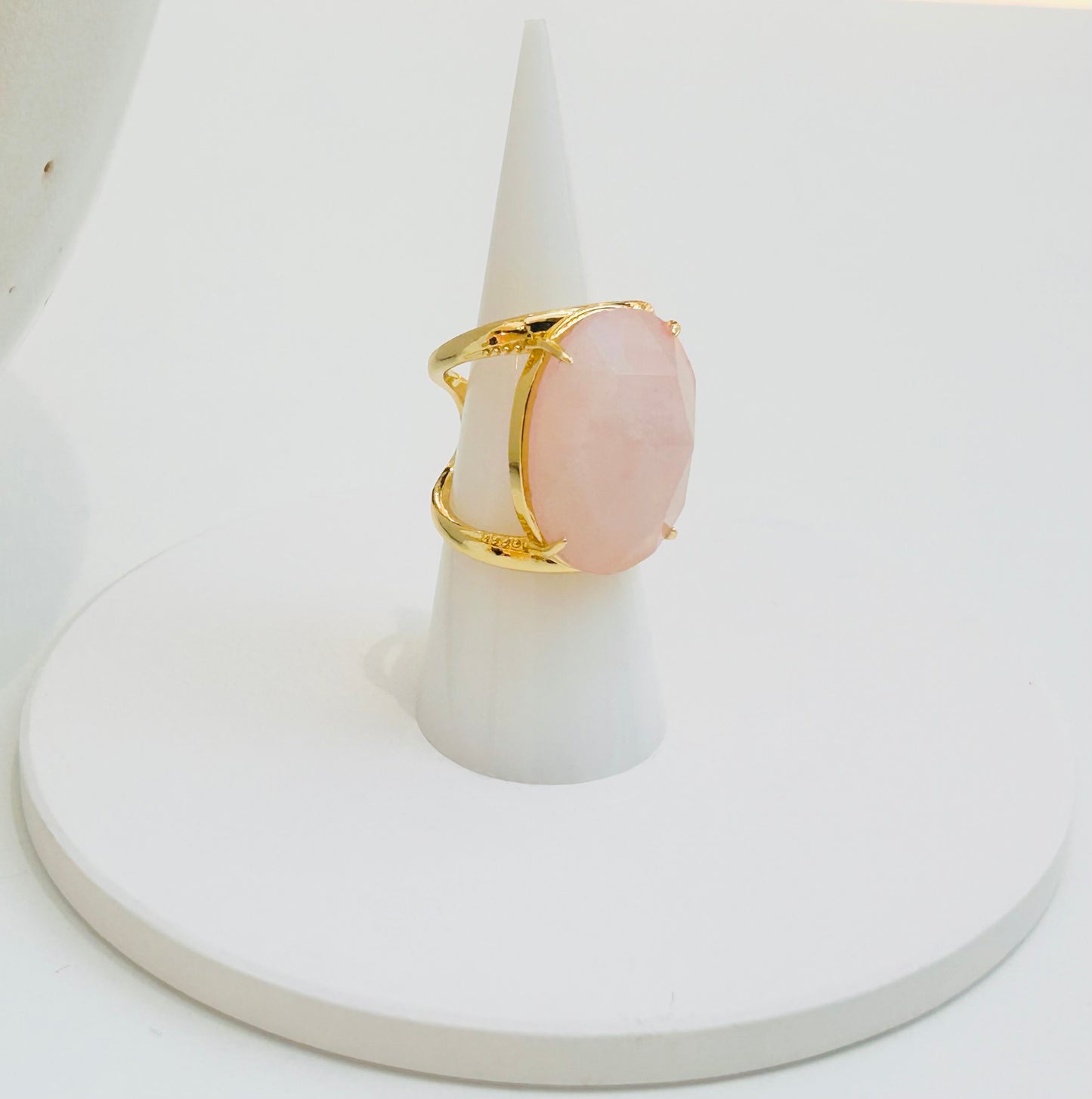 
                  
                    A gold ring with a large oval pale pink rose quartz stone, displayed on a white cone-shaped ring holder over a round white base.
                  
                