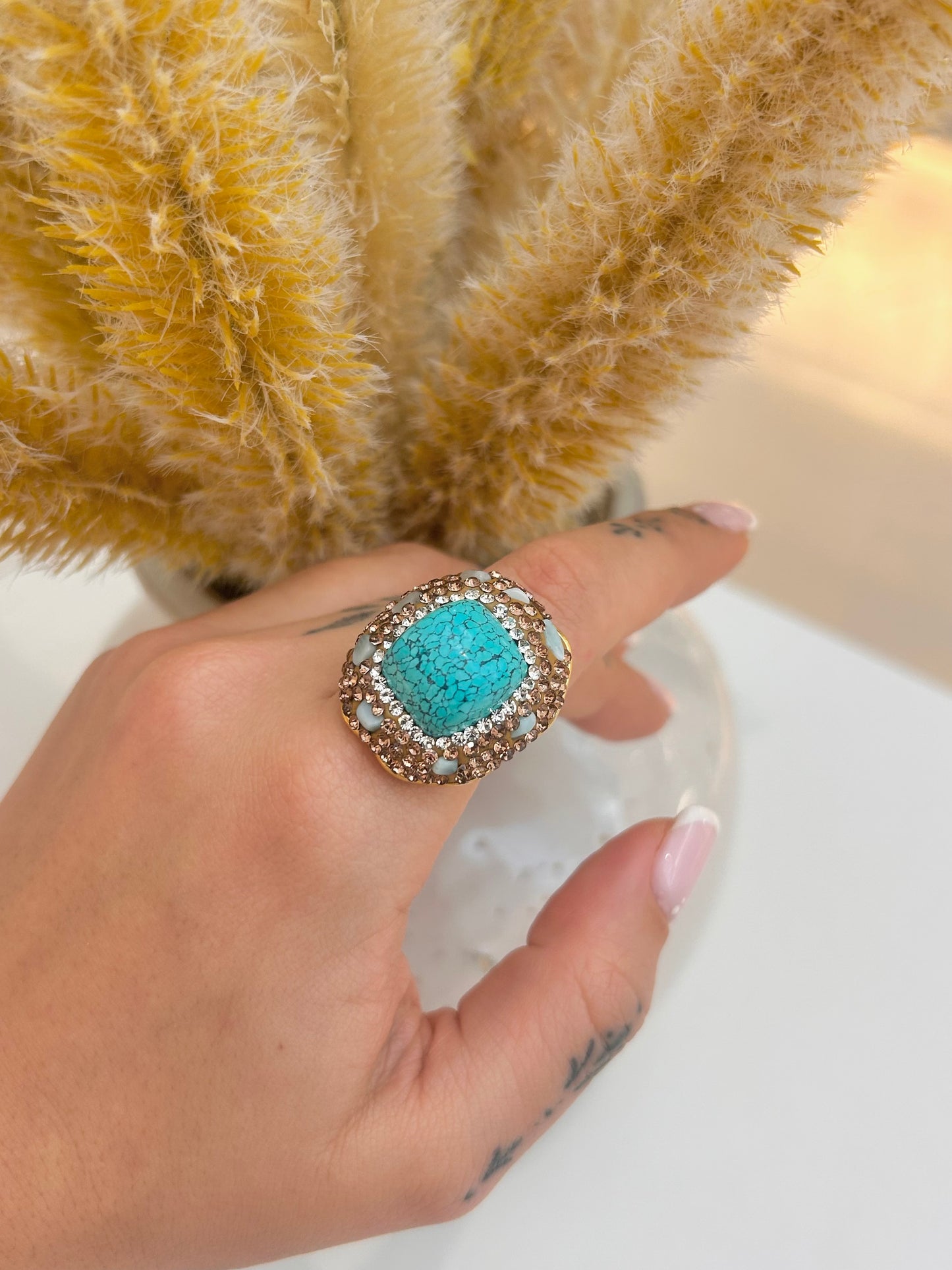A close-up of a hand wearing a large statement ring with a square turquoise center and a halo of small clear stones, set against a background of dried pampas grass.