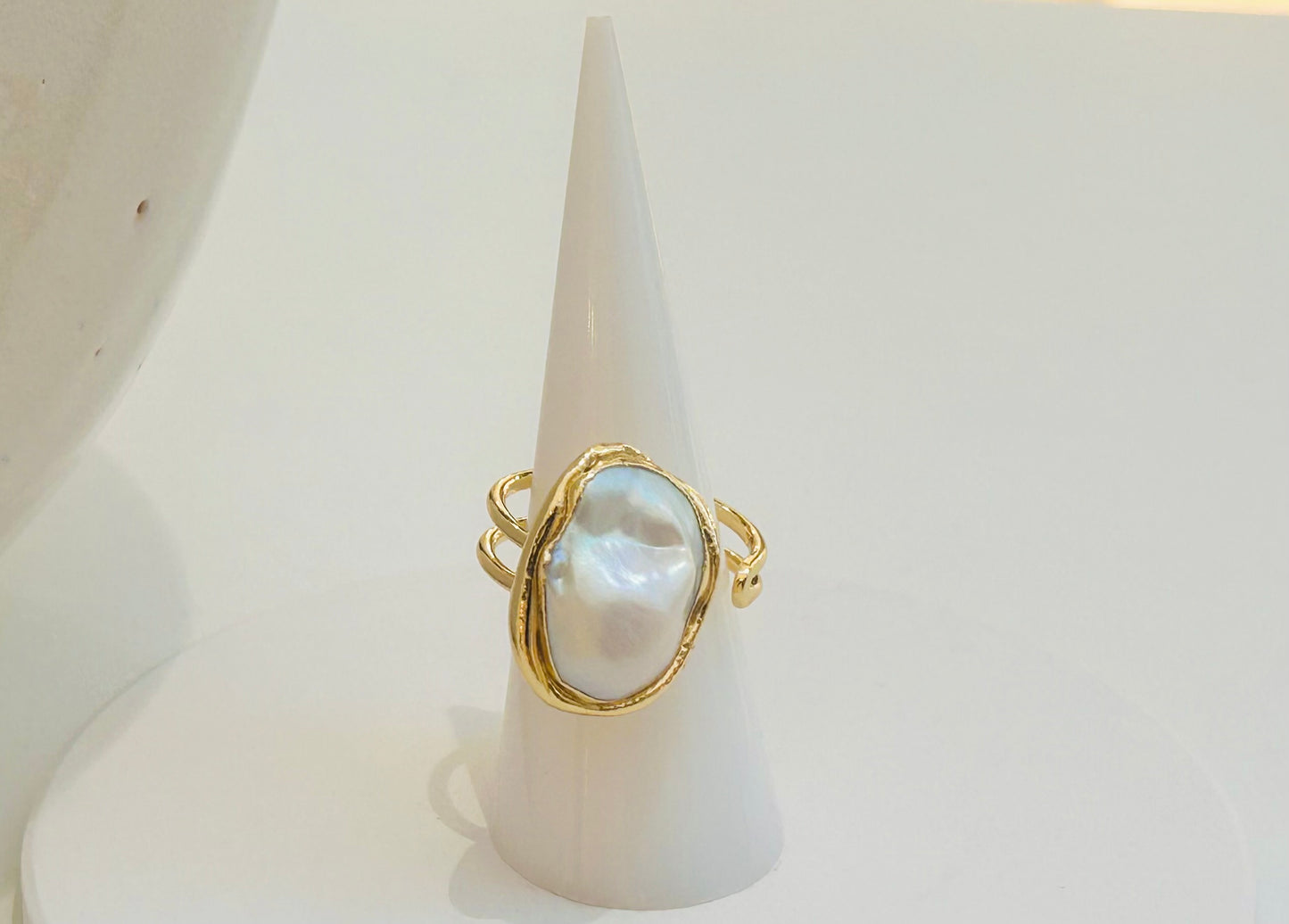 
                  
                    A gold ring with a large, irregular white baroque pearl set in an oval bezel, displayed on a white cone jewelry stand against a clean white background.
                  
                