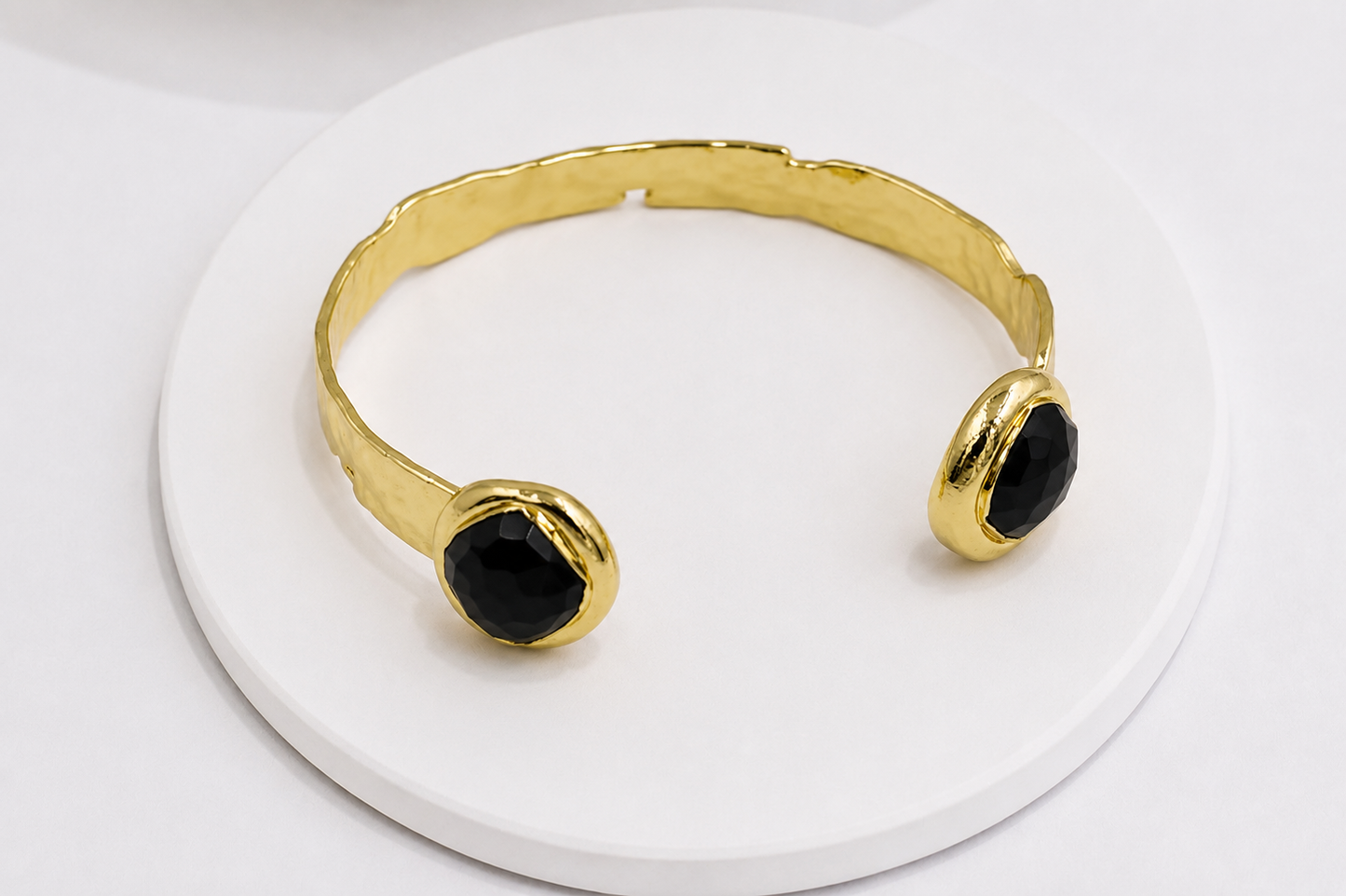 Gold cuff bracelet with two oval black obsidian stone settings, displayed on a round white pedestal.
