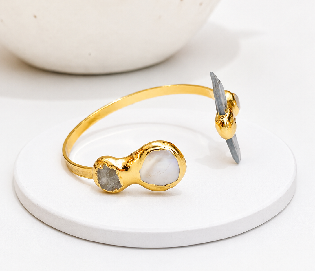 A gold-plated open cuff bracelet with a toggle clasp and two bezel-set cabochon stones, displayed on a white round stand against a white background.
