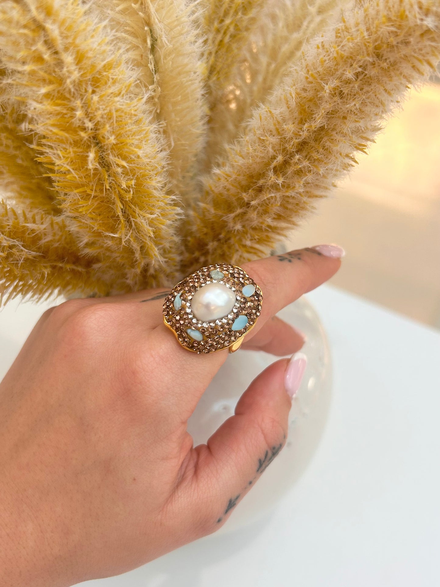 A gold ring with a large white pearl center and light anmazonite accents, worn on a hand with a tattoo, positioned in front of dried pampas grass.