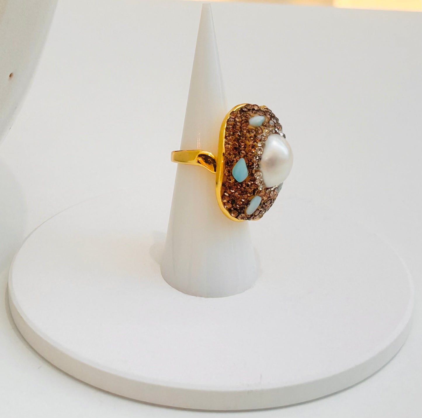 
                  
                    A gold ring with a large white pearl and brown textured stones accented by light amazonite stones, displayed on a white cone stand.
                  
                