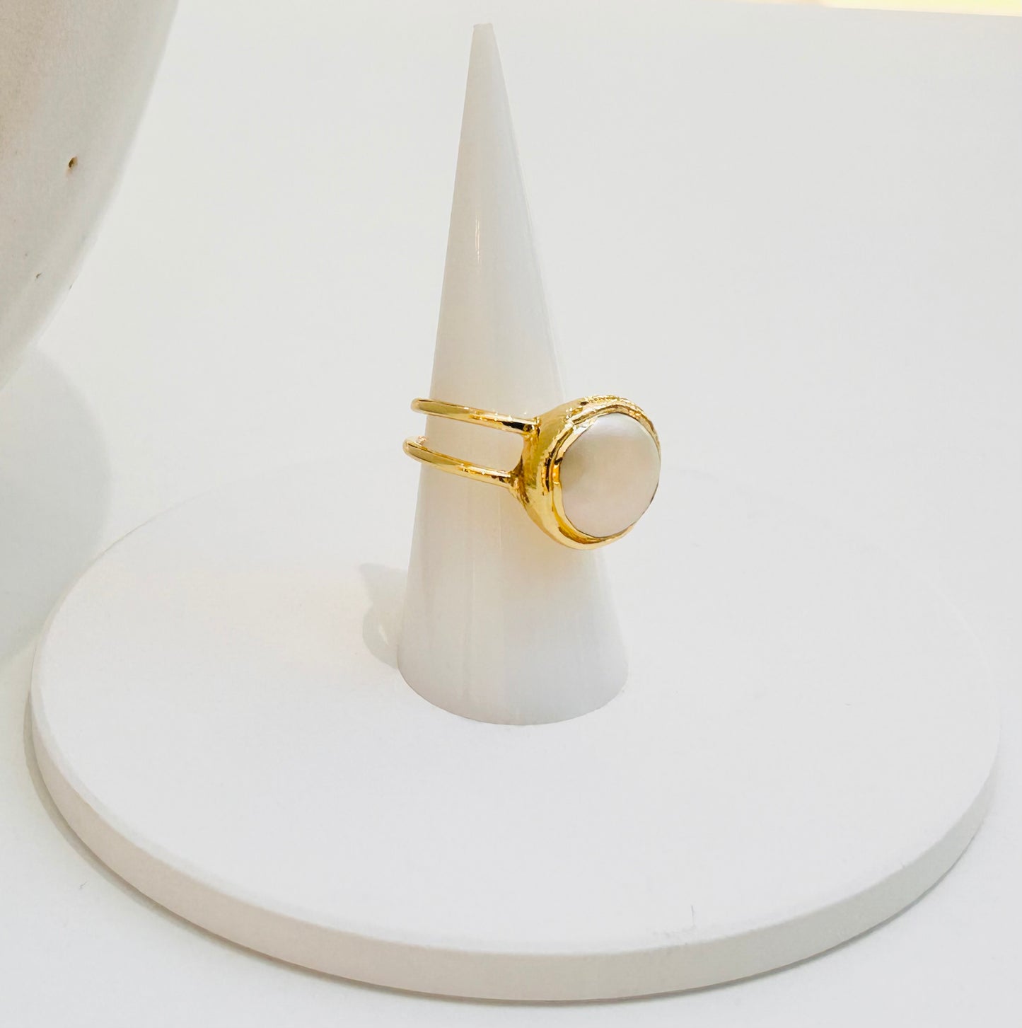 
                  
                    Elara Freshwater Pearl Statement Ring displayed on a white jewellery stand, featuring a round natural pearl in a textured double-band gold-plated design.
                  
                