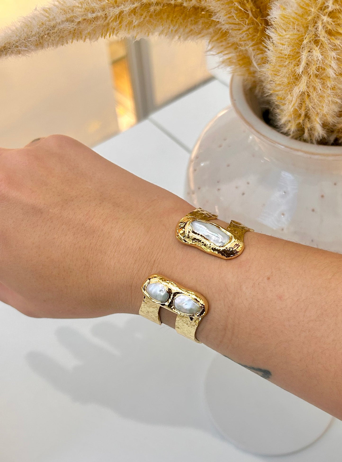 A close-up of a person's forearm wearing a gold cuff bangle with white freshwater pearl rectangular inlays, set against a white tabletop with a ceramic vase and dried yellow stems in the background.