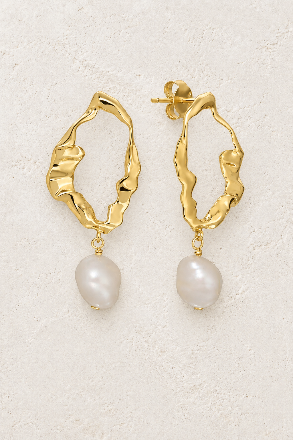 Gold organic hoop earrings with freshwater pearl drops on a warm off-white textured plaster background
