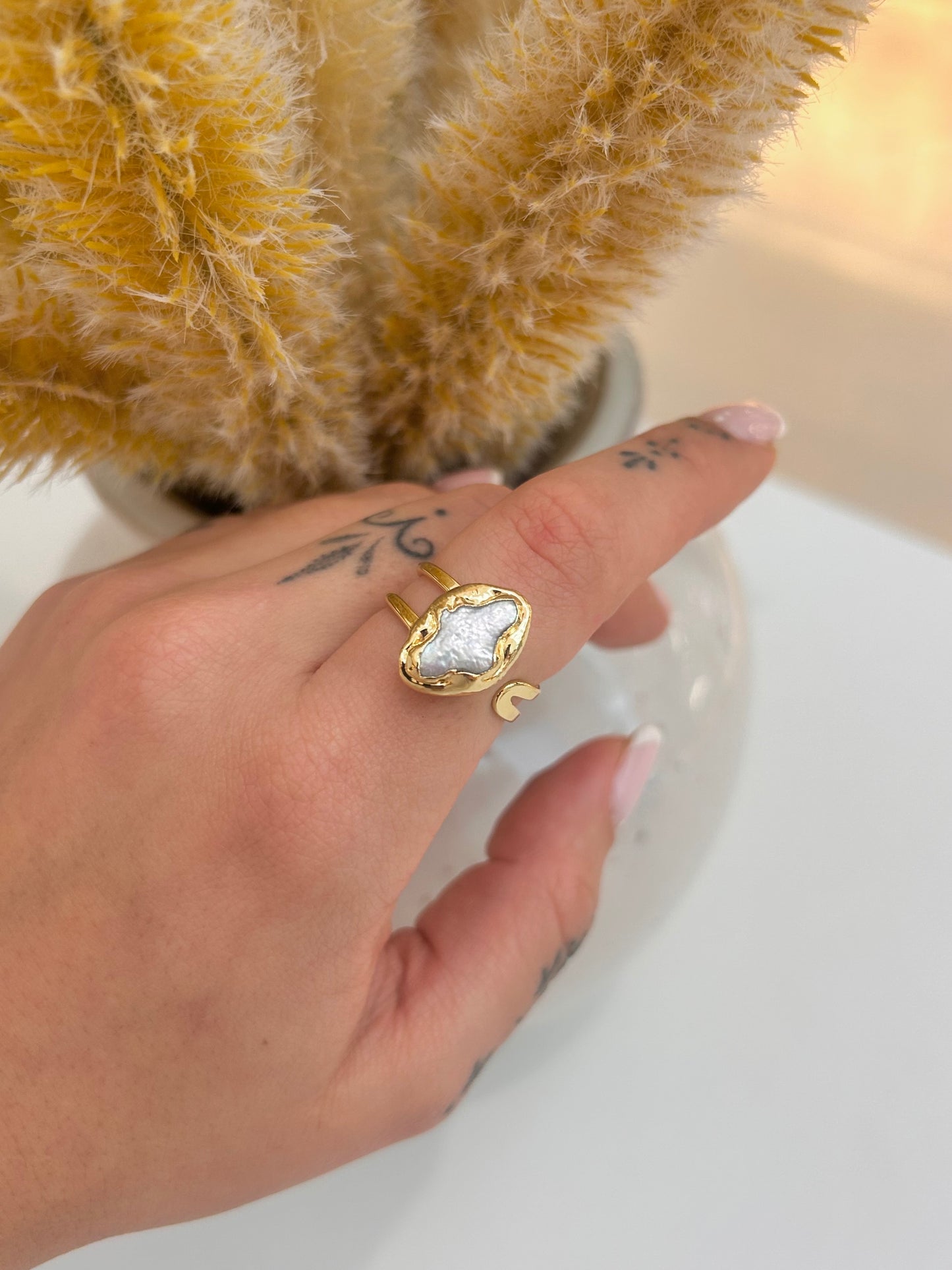 A gold ring with a large, irregular white pearl-like stone is shown worn on a person's finger, with a soft-focus background featuring a yellow textured plant and a white surface.