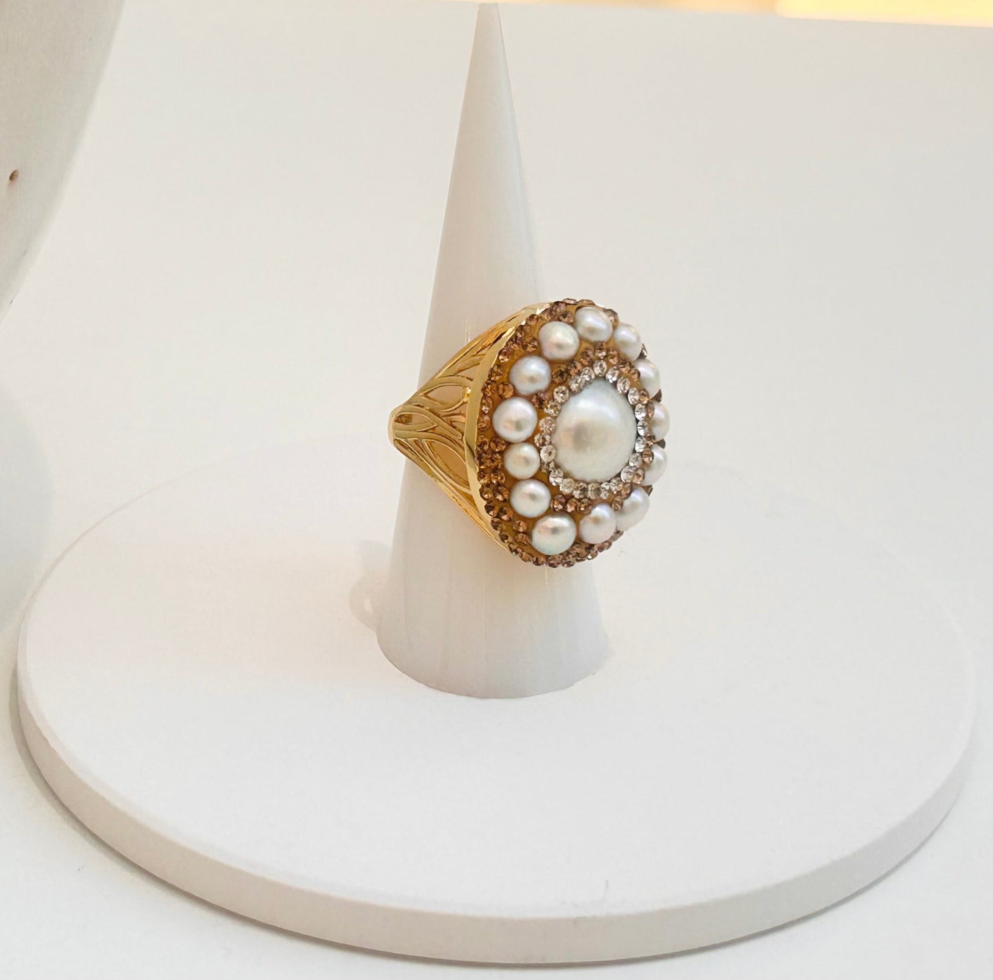 
                  
                    A vintage-style gold ring with a large white pearl center and a halo of smaller pearls, displayed on a white cone stand against a white background.
                  
                