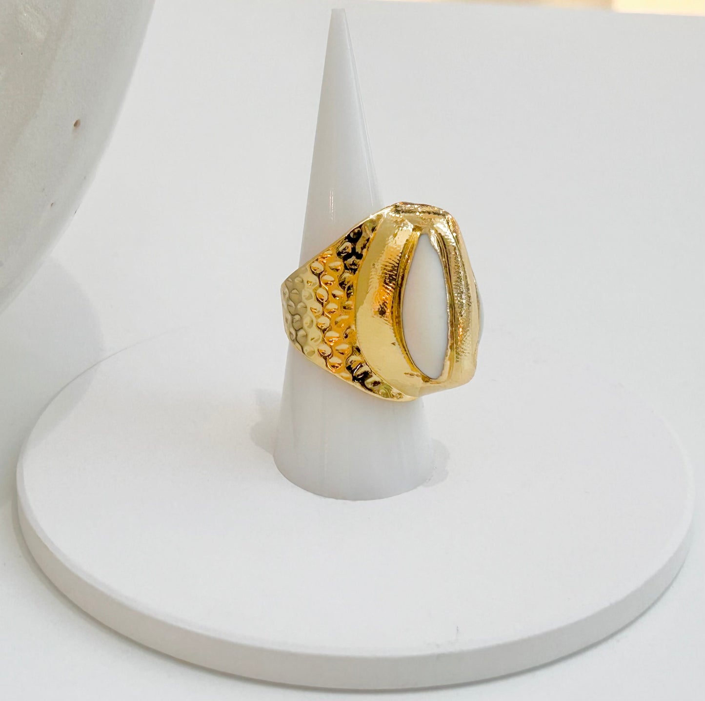
                  
                    Marella Cowrie Shell Statement Ring displayed on a white jewellery stand, featuring a natural white cowrie shell in a hammered gold-plated setting.
                  
                
