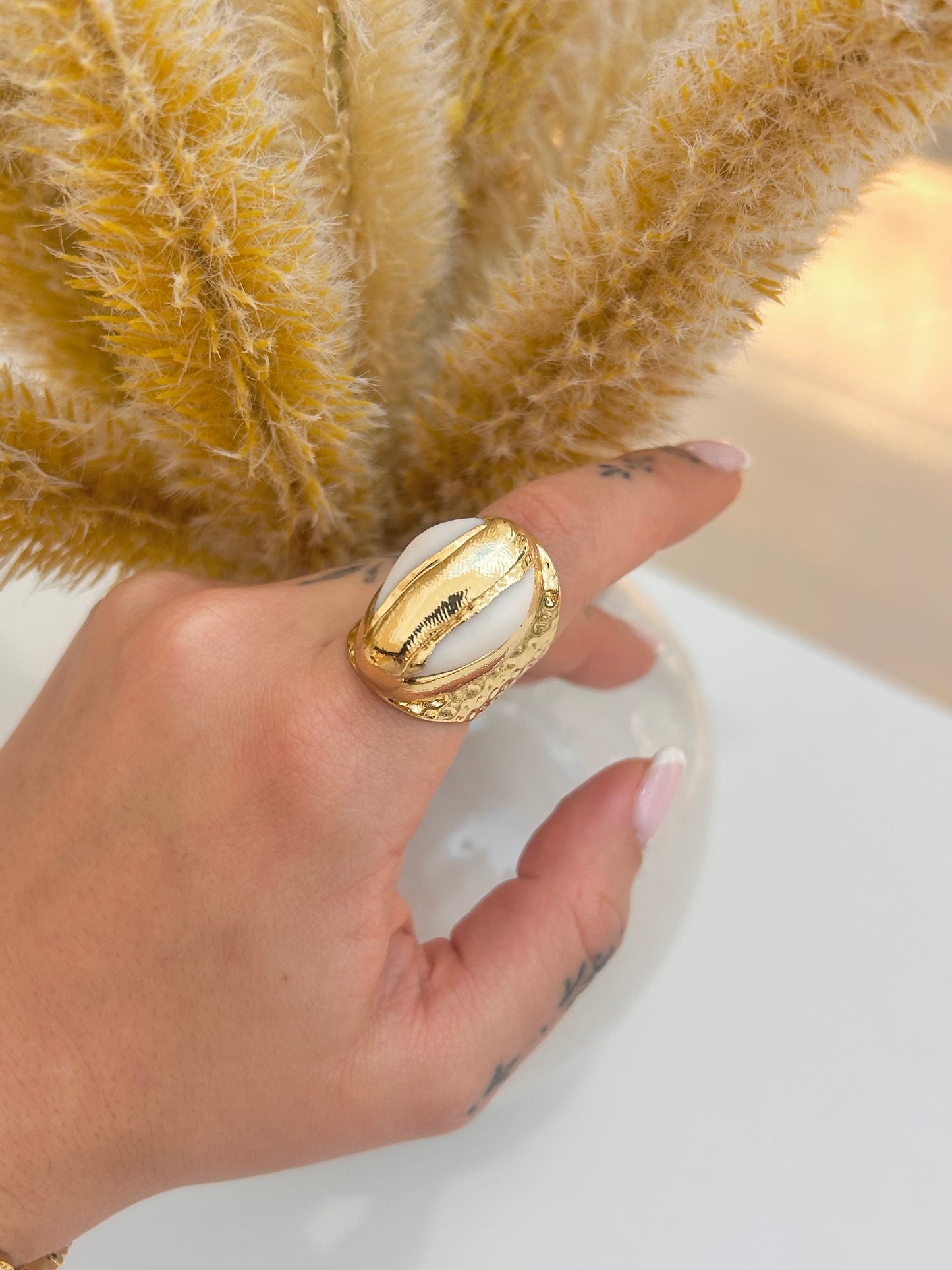 Hand wearing the Marella Cowrie Shell Statement Ring featuring a white cowrie shell and textured hammered gold-plated band.