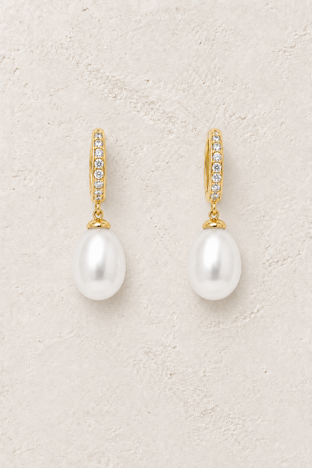 Pearl Lumiere crystal huggie earrings with freshwater pearl drops on a warm off-white textured background