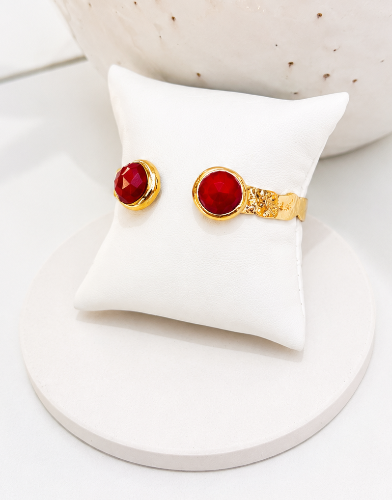 
                  
                    A gold plated open cuff bangle with round red jade stones, displayed on a small white cushion.
                  
                