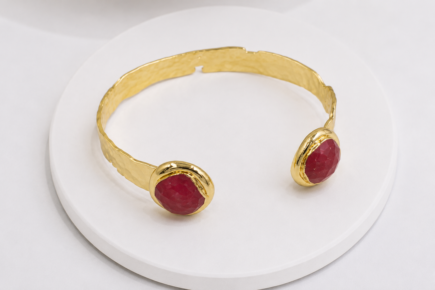 A gold cuff bracelet with two oval red jade gemstone accents, displayed on a white circular stand against a white background.