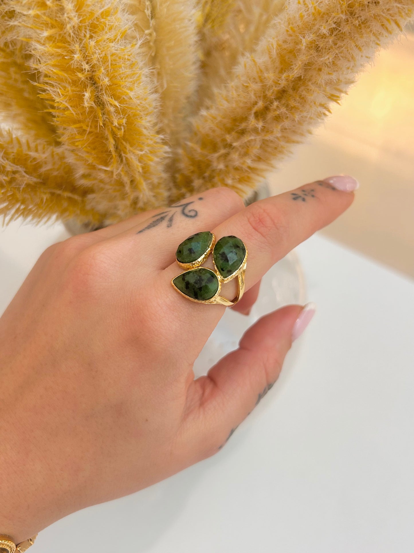 A gold ring with three marquise-cut African turquoise gemstones worn on a person's finger, set against a background of a yellow fluffy vase and a white surface.