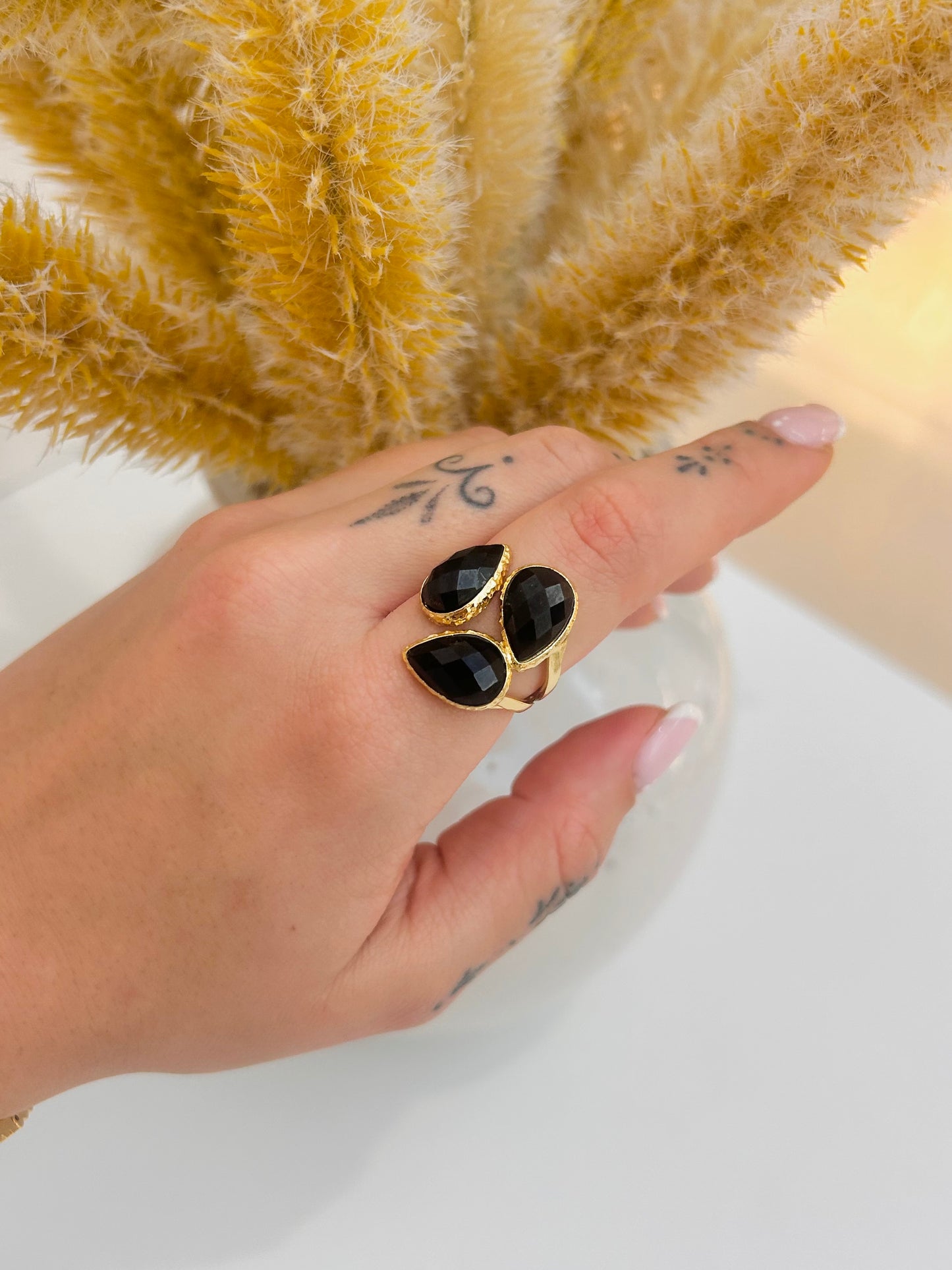 A gold ring with three faceted black obsidian teardrop gemstones worn on a person's hand, with dried pampas grass in the background.