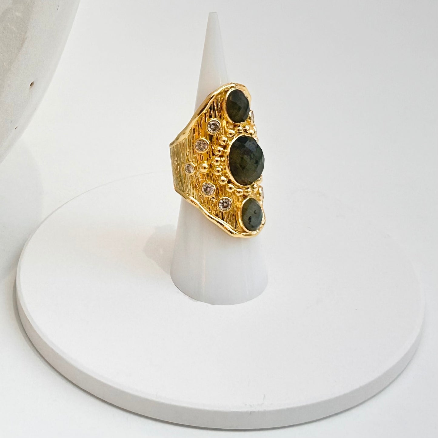 
                  
                    A gold ring with three large oval dark labradorite stones and small clear accent stones, displayed on a white cone stand against a white background.
                  
                