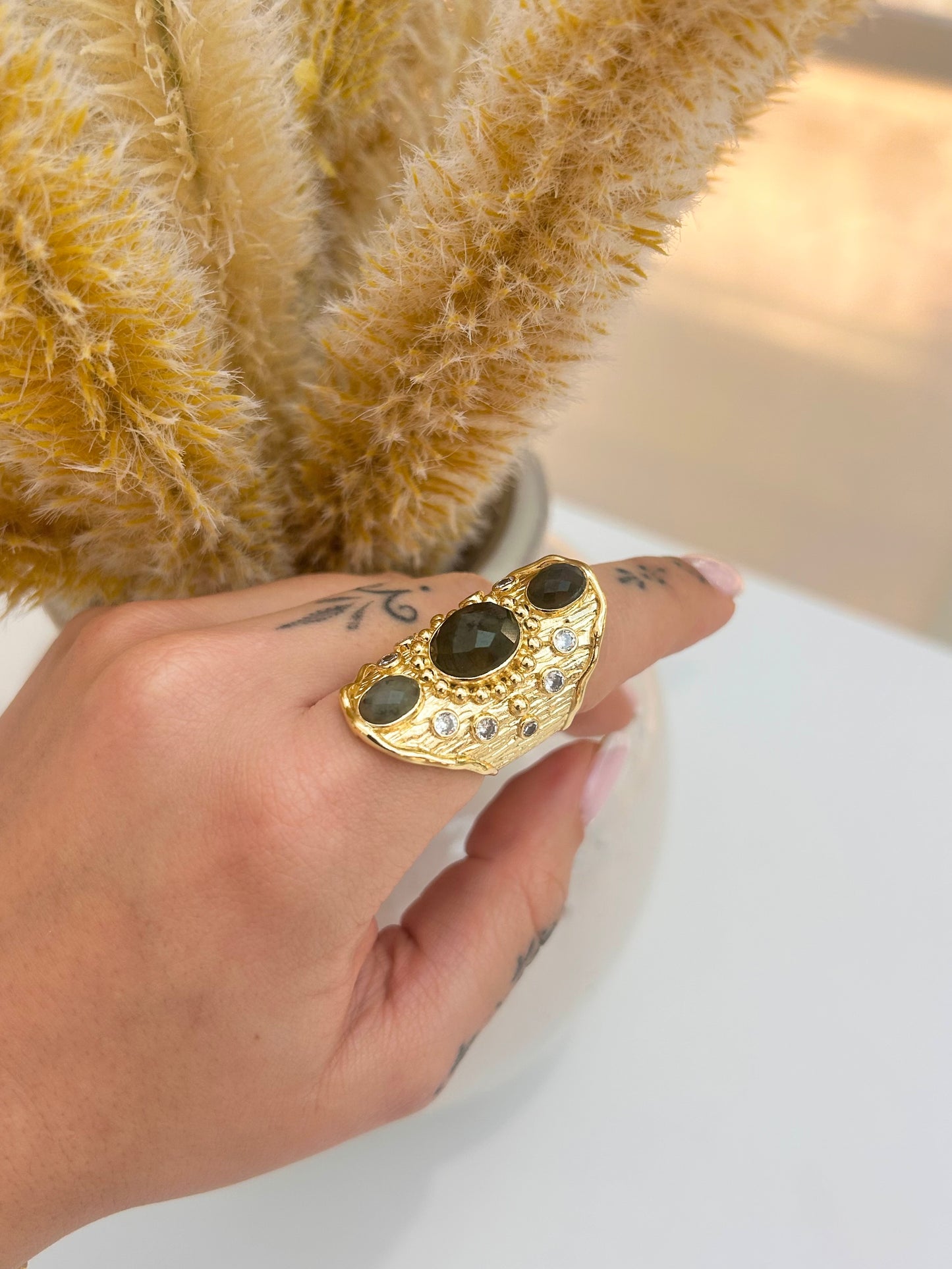 A gold ring with three labradroite gemstones and small clear accent stones, worn on a hand with a tattoo, styled with dried pampas grass in a white pot.
