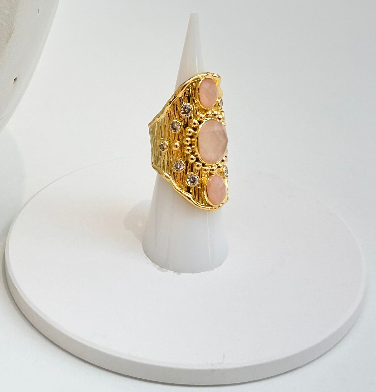 
                  
                    A gold ring with three oval pale pink rose quartz stones and small clear accent stones, displayed on a white cone stand against a white background.
                  
                