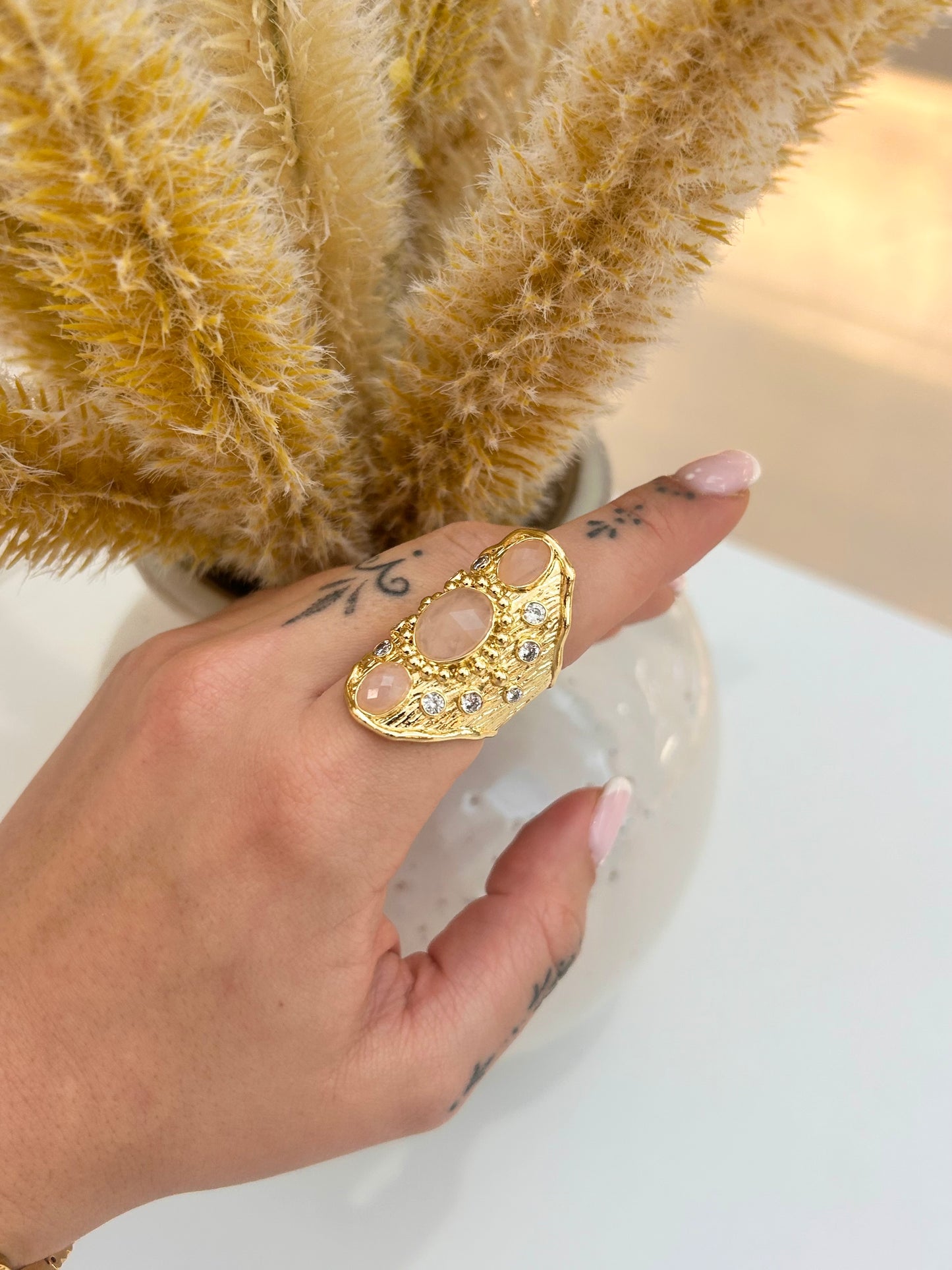 A gold crescent moon ring with a central oval pink rose quartz stone and small clear stones, worn on a person's finger. The hand is positioned in front of a vase with dried pampas grass on a white surface.