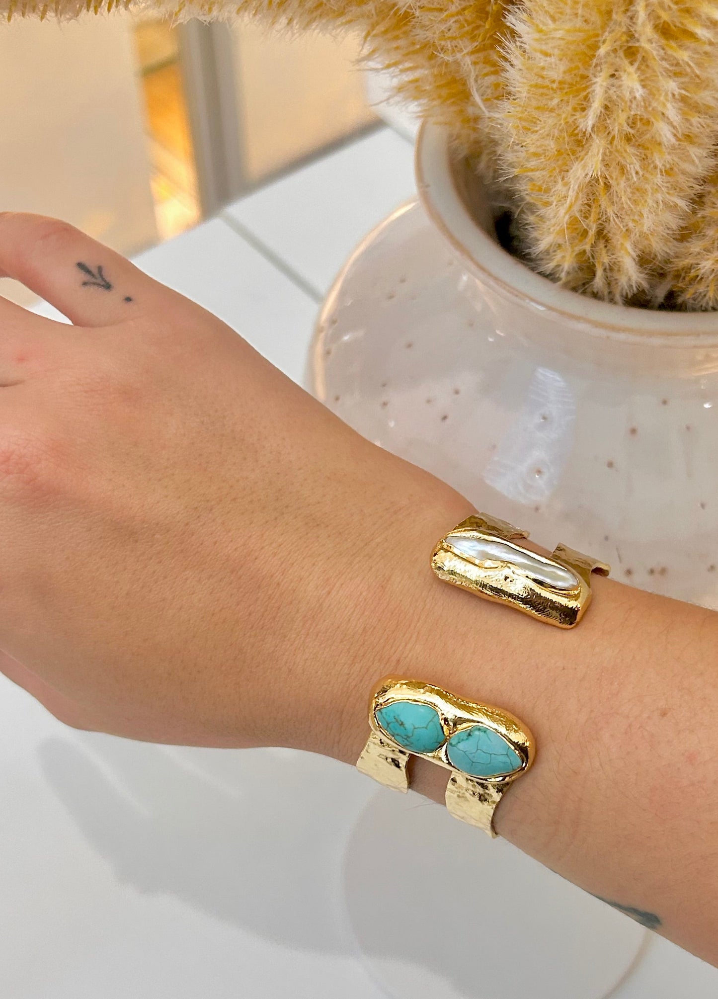 A gold cuff bracelet with two oval turquoise stones one side and freshwater pearl on the other, worn on a person's wrist, with a white speckled vase and a beige plush toy in the background.