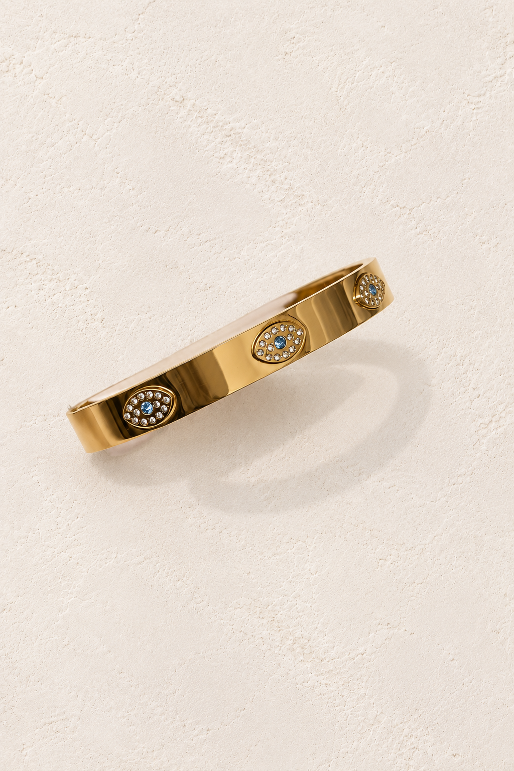 
                  
                    Zaria Gold Evil Eye Bangle on cream beige textured background
                  
                